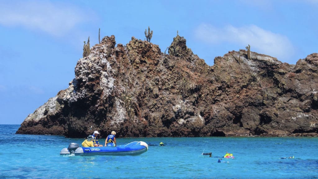 A lone snorkeler swims in brilliant blue water near a small boat, with volcanic cliffs in the background.