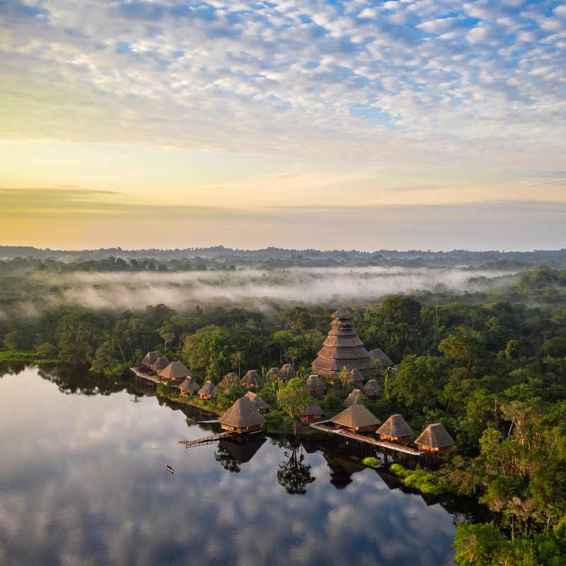 Aerial view of Napo Wildlife Center's thatched-roof lodge on a misty Amazon lagoon in Ecuador at dawn.